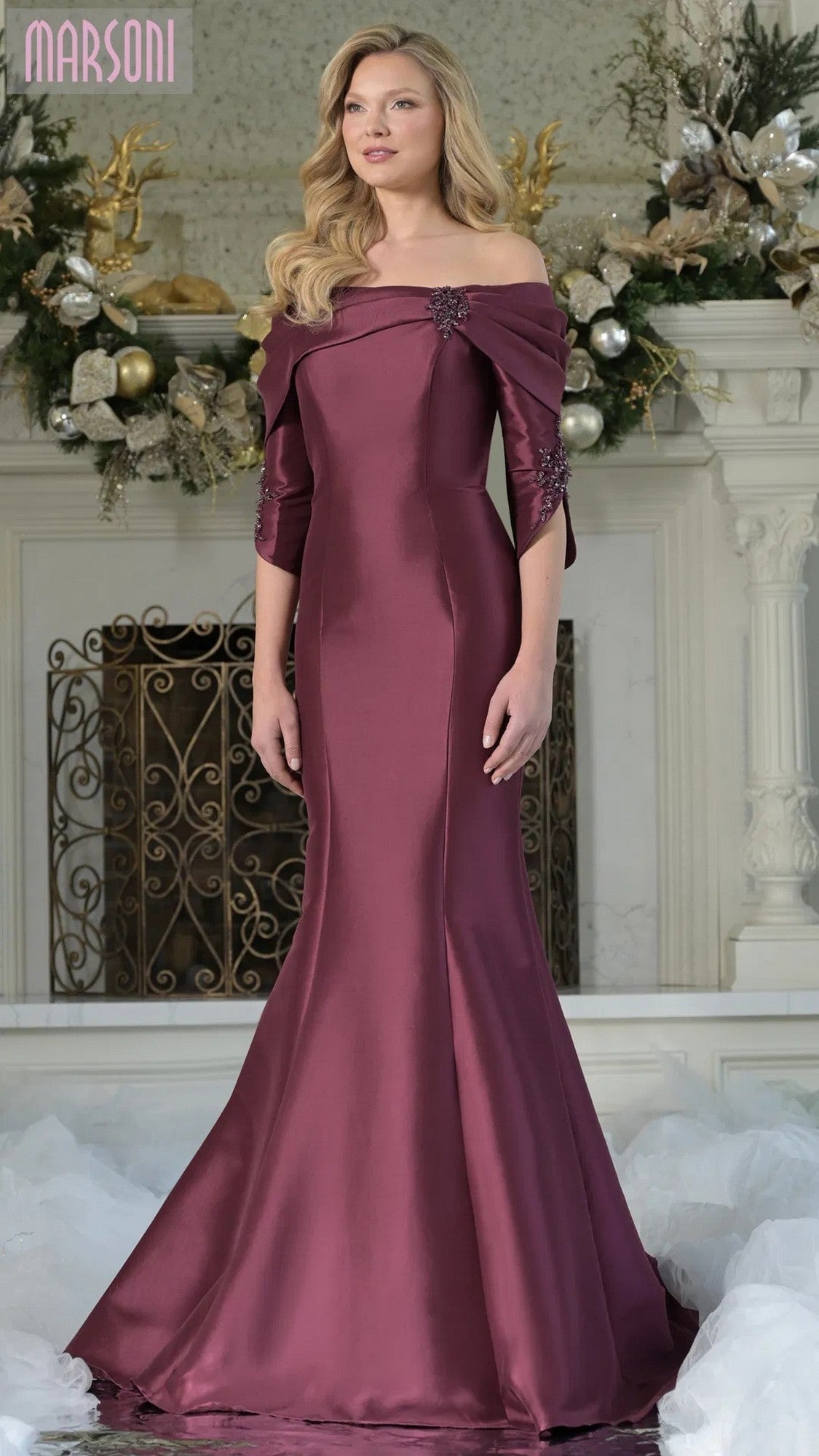 A woman stands indoors wearing the Marsoni MV1360 long formal dress in wine, an off-the-shoulder, floor-length mermaid gown with three-quarter sleeves and a detailed bodice. Holiday decorations can be seen in the background.